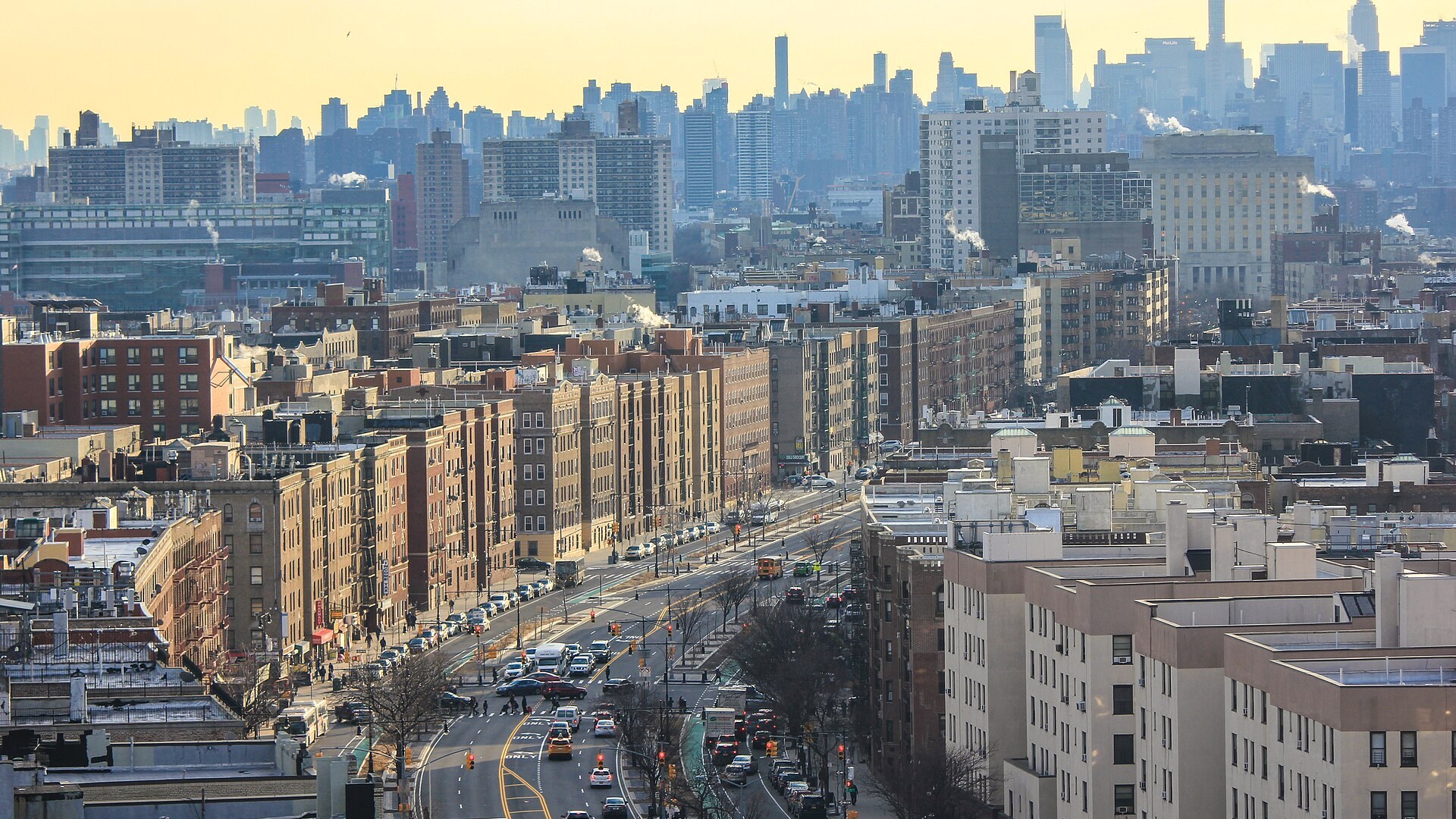 Rows of prewar apartment houses along the Grand Concourse in the Bronx at dusk, with the Manhattan skyline behind them.